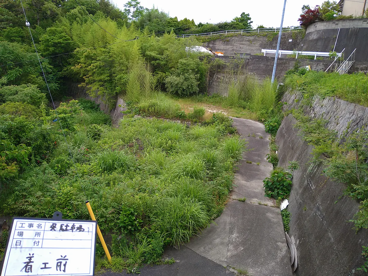 施工前：草が茂った敷地。奥に擁壁と建物。「着工前」の標識が立っている